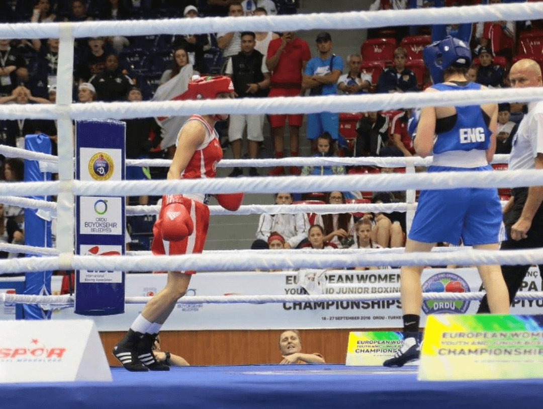 Emma Dolan at the European Championships wearing a blue England vest in the ring. Both boxers an in their corners, preparing for the round to start.