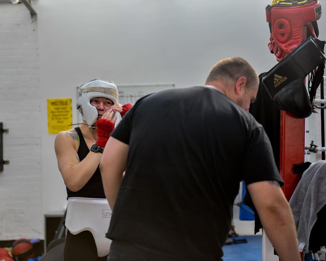 Amy Greatorex putting on her headguard. Her coach is stood in the foreground with his back to the camera, he's leaning as if to pick something up.