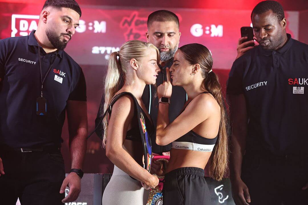 Tysie Gallagher and Tori-Ellis Willetts face-off onstage. Tysie is wearing her British and Commonwealth titles on either shoulder and Tori is gesturing that she will silence Tysie, with her index finger on her lips.